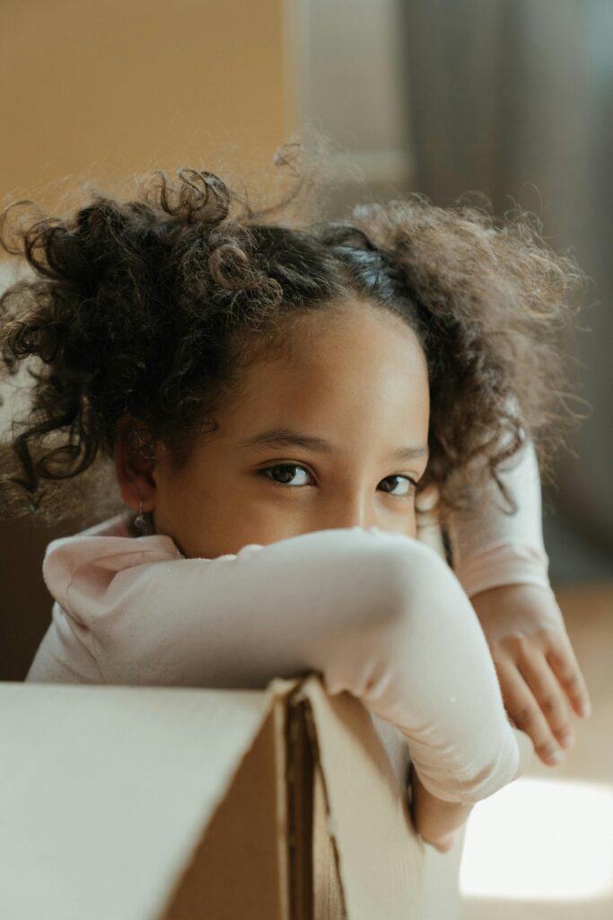 Cute child with curly hair peeks from inside a cardboard box, symbolizing moving or playtime.